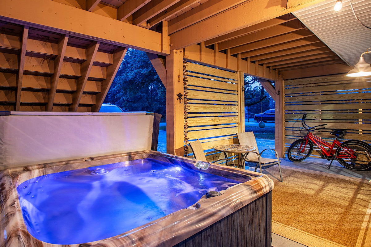 A private hot tub with illuminated water is situated under a wooden structure, surrounded by a rustic vibe. A bike is parked nearby, alongside two simple chairs. The warm lighting and natural wood enhance the relaxing atmosphere.