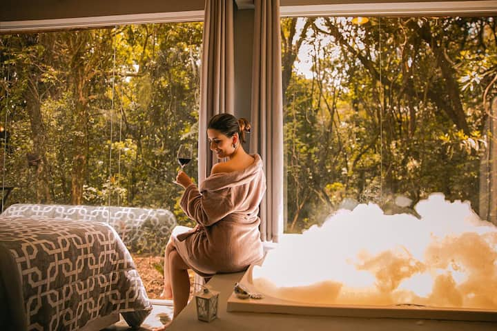 Moça sentada na beira da banheira em casa com vidro e vista da natureza