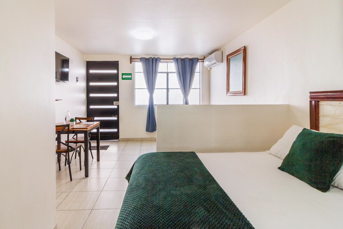 A modern loft is displayed, featuring a spacious bed adorned with a textured green blanket. A wooden table with two chairs is positioned nearby under a large window framed by blue curtains. A wall-mounted television and an air conditioning unit are visible.