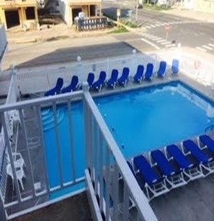 A clear view of the inviting pool area is showcased, surrounded by lounge chairs arranged in neat rows. The pool's vibrant blue water reflects the sunlight, providing a refreshing retreat. Nearby, an intersection and other buildings can be seen, completing the setting.