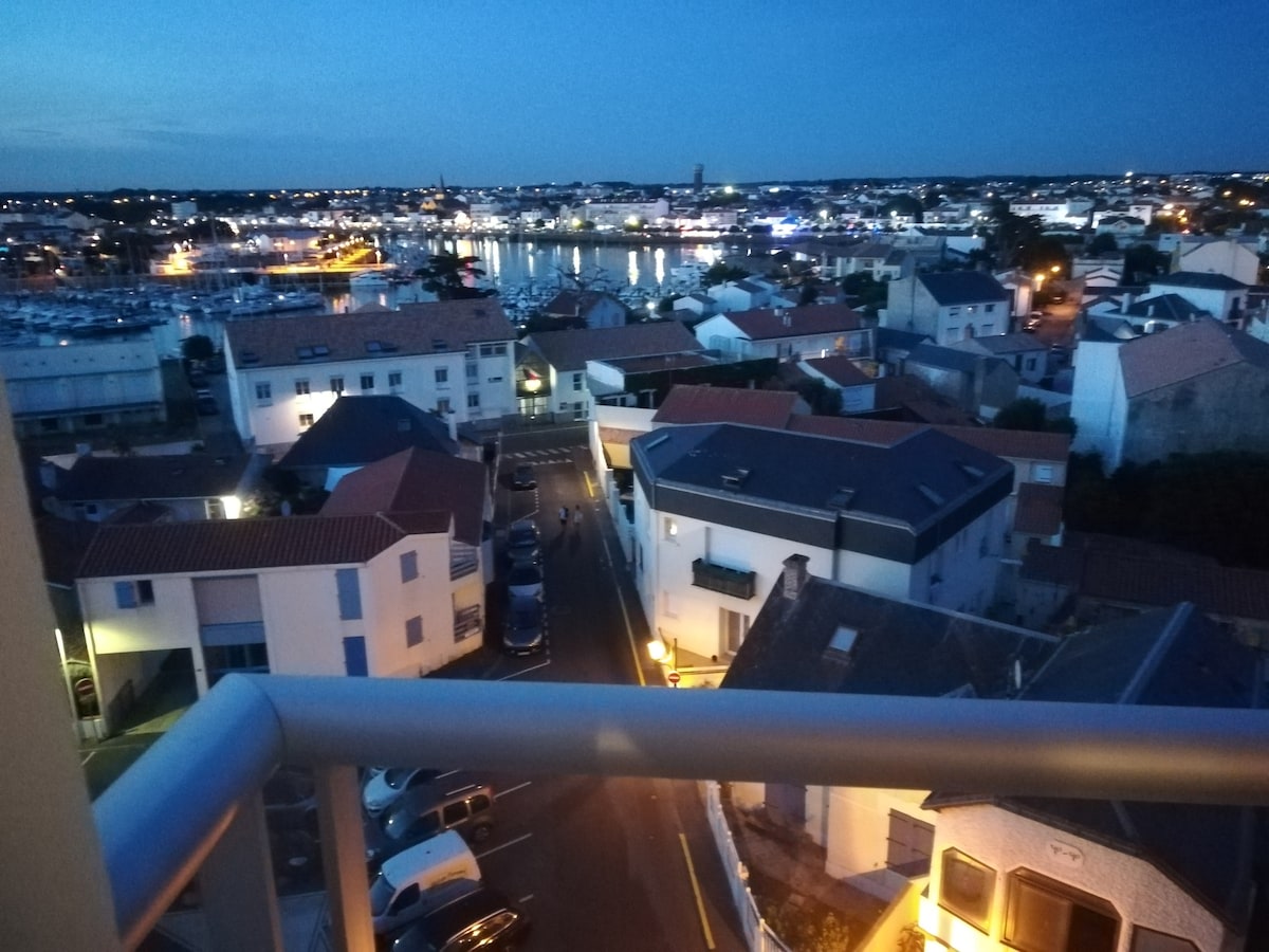 A nighttime view shows the marina and waterfront, illuminated by soft lights reflecting on the water. The sight includes a mix of rooftops and streets below, with some buildings displaying warm, inviting glows from illuminated windows.