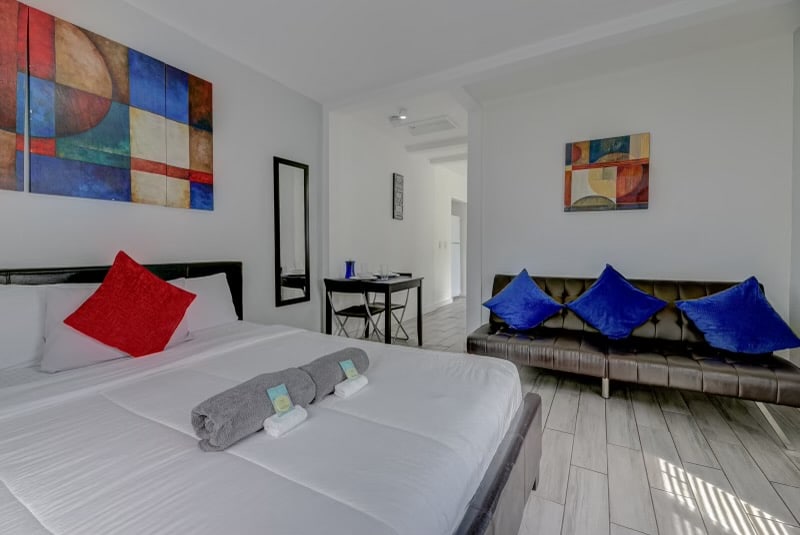 A spacious sleeping area features a large bed with a white comforter and colorful decorative pillows. Bold artwork adorns the walls, while a sleek mirror reflects the room. A brown sofa with blue accent pillows and a small dining table with chairs complete the inviting layout.