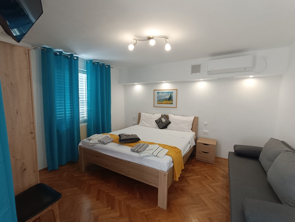 A well-lit sleeping area features a spacious bed covered with fresh linens and accented by a bright yellow throw. A comfortable seating area with a dark sofa is present, along with a bedside table. Soft blue curtains adorn the window, allowing natural light to fill the room.