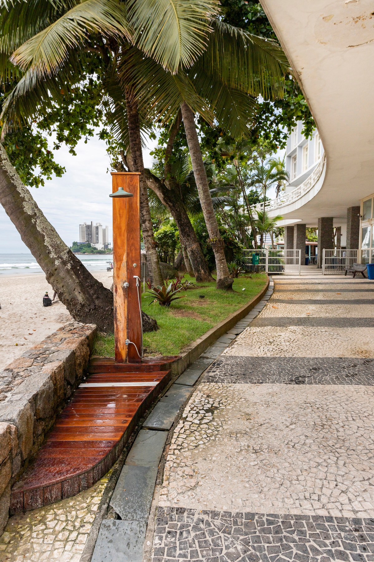 An outdoor shower is seen made of wood, positioned alongside a walkway lined with palm trees and greenery. The pathway, featuring a mix of stone and tile, leads towards the sandy beach, providing easy access to the ocean.