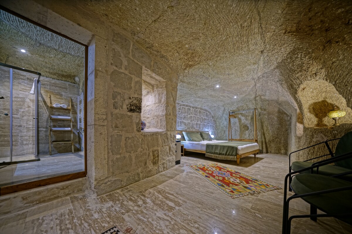 A cave room features natural stone walls and a spacious bed adorned with neutral linens. A colorful rug lies on the floor, adding a pop of color. A modern shower area and a staircase are visible, enhancing the room's unique design.