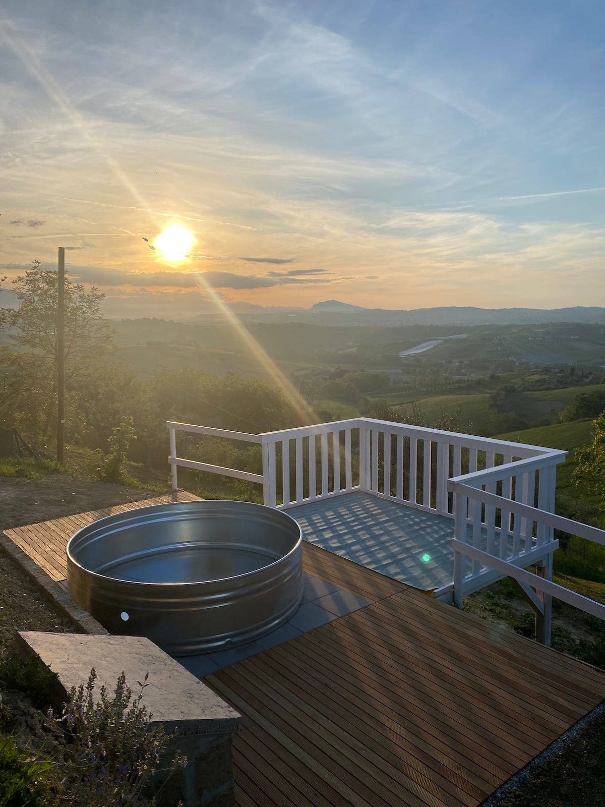 An outdoor terrace features a circular hot tub, overlooking a serene landscape. Sunlight streams through the sky, casting a warm glow over rolling hills and distant mountains. The wooden deck complements the natural surroundings, creating a tranquil atmosphere for relaxation.
