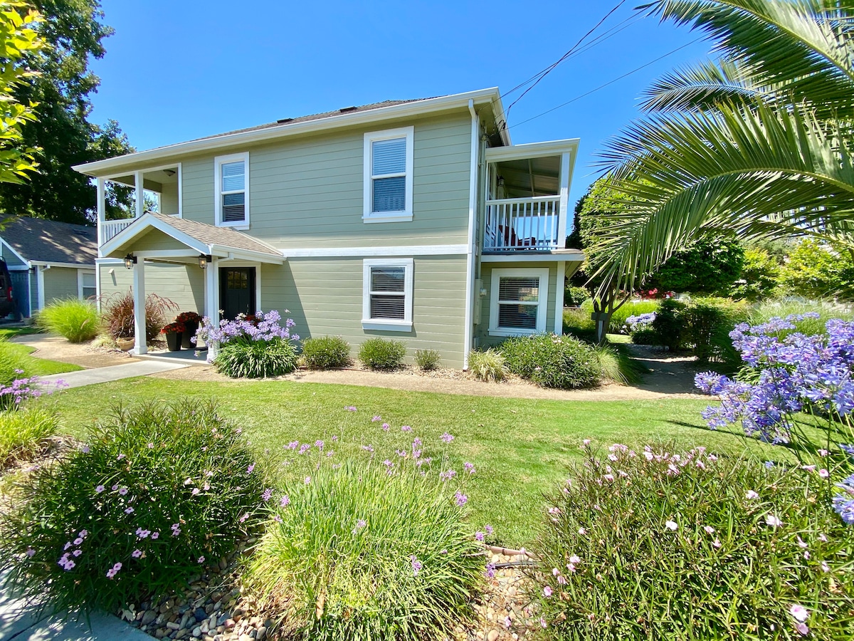A two-story light green house is set amidst lush landscaping, featuring well-maintained lawns and flowering bushes. A covered porch is visible on the upper floor, and trees provide shade around the property. The clear blue sky adds to the inviting outdoor ambiance.