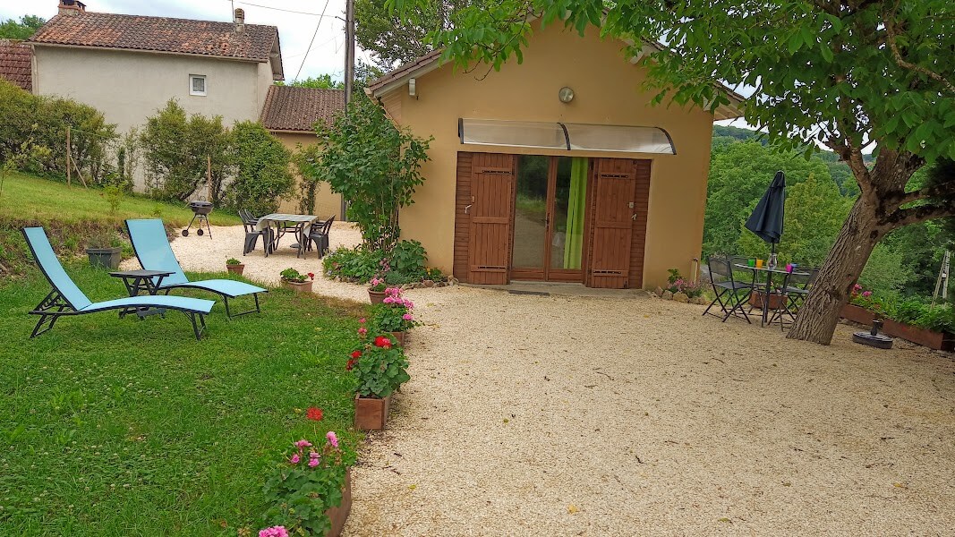 A charming outdoor area features a gravel path leading to a cozy gîte. Two light blue loungers are positioned on the grass, alongside potted flowers. A dining table with chairs and an umbrella are set up, inviting relaxation amid the greenery.