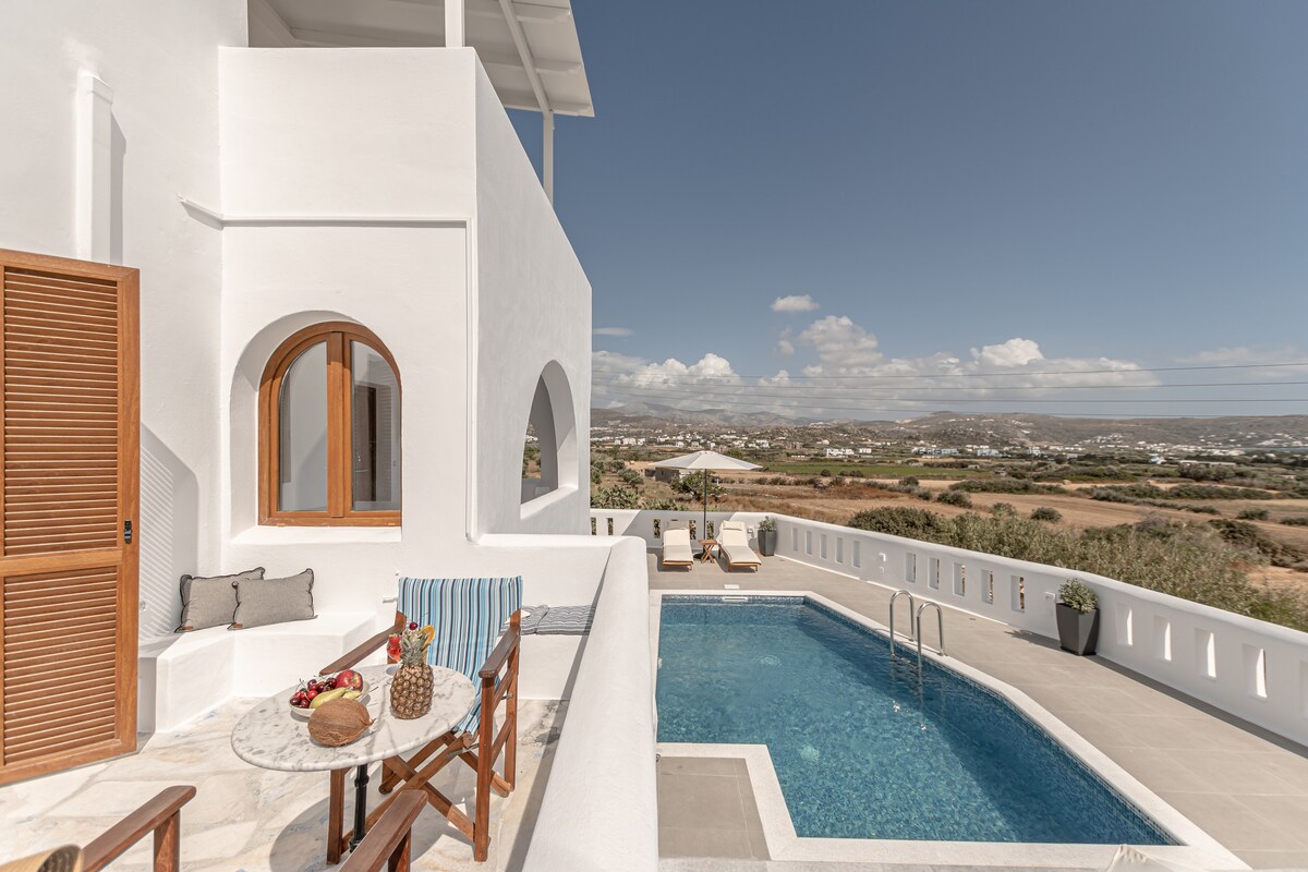 A white-washed exterior features arched windows and a balcony overlooking a pool. The pool area includes lounge chairs alongside a small table adorned with a basket. Scenic views of the surrounding landscape are visible in the background, enhancing the outdoor space.