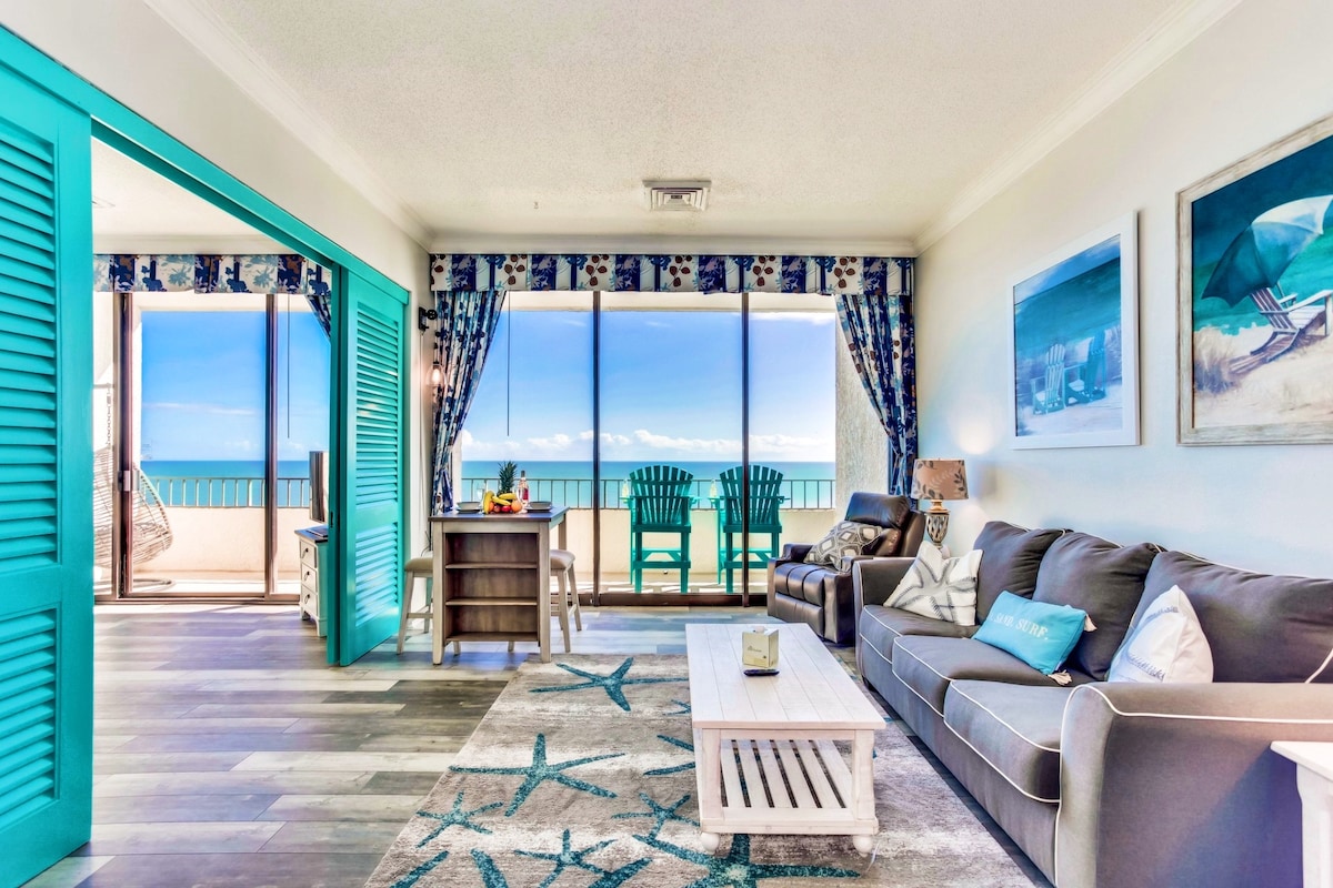 A spacious living area is highlighted by large windows offering ocean views. The room features a comfortable sofa and a coffee table, with a dining area in the background. Bright turquoise accents are complemented by nautical-themed decor, creating a relaxed ambiance.