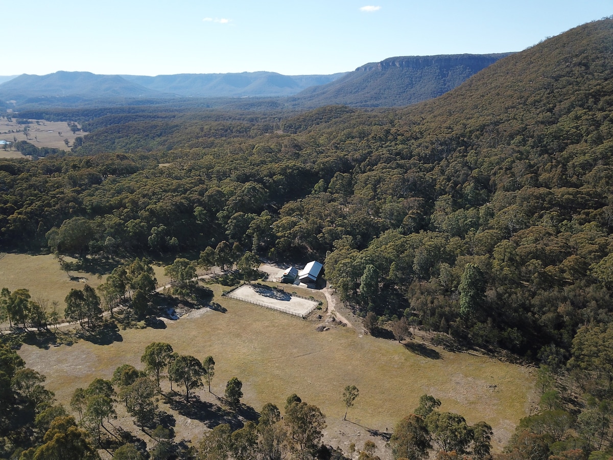An aerial view captures the cottage nestled among lush greenery and rolling hills, surrounded by expansive cleared land. The distant mountain range provides a serene backdrop, while the open space invites exploration of the surrounding natural beauty.