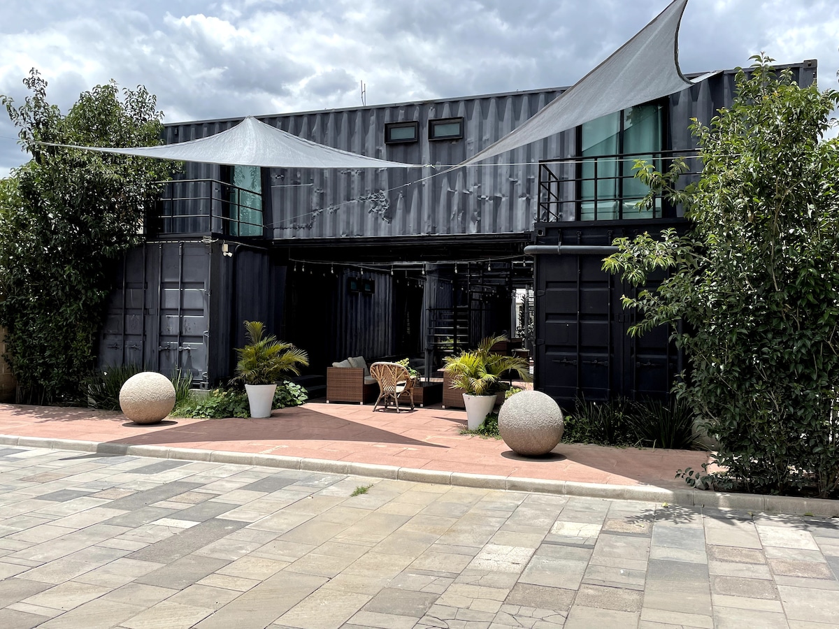 A modern loft design is presented with an industrial aesthetic, featuring dark shipping containers accented by greenery. Shade sails stretch overhead, providing shelter to a seating area. Large planters are positioned alongside smooth stone walkways, enhancing the welcoming entrance to the space.