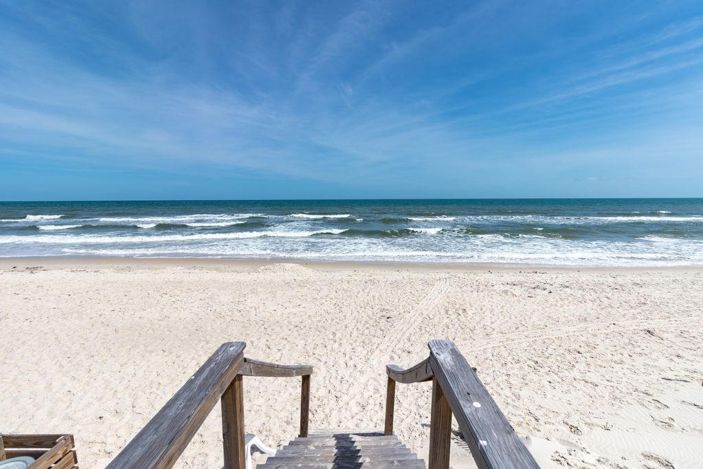 A set of wooden steps leads down to a sandy beach, where gentle waves lap at the shore. The expansive ocean stretches into the horizon under a clear blue sky, creating an inviting coastal atmosphere.