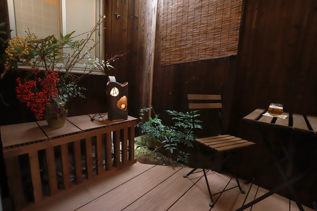 A peaceful outdoor space features wooden decking with a small bistro table and folding chair. Lush greenery surrounds the area, complemented by a decorative lantern and vibrant plants. Natural light filters in, creating a serene atmosphere for relaxation.