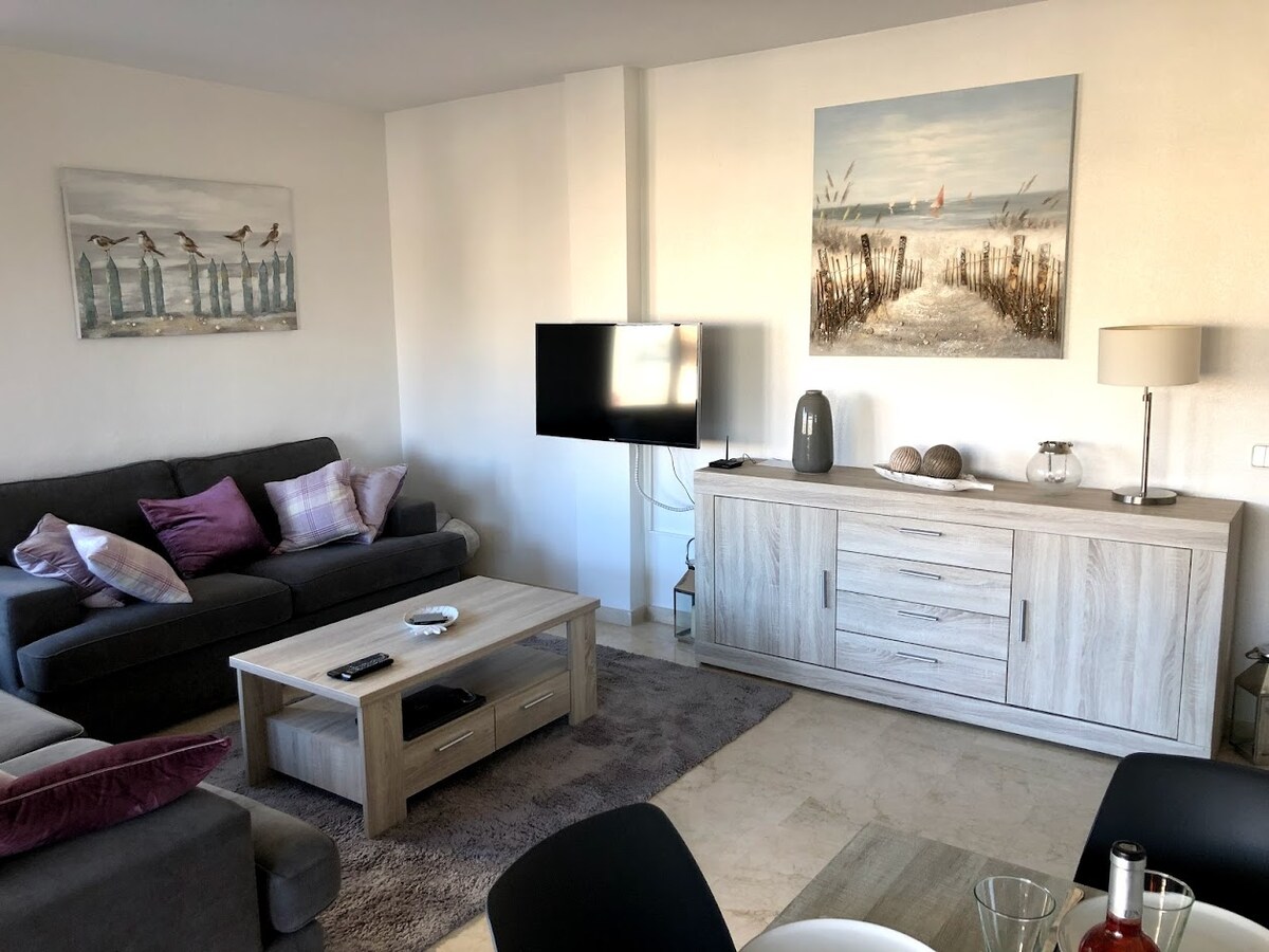 A spacious living area is presented, featuring a comfortable gray sofa adorned with decorative pillows. A flat-screen TV is mounted on the wall across from the seating. A light wooden sideboard holds decorative items, complemented by a soft area rug on the floor.