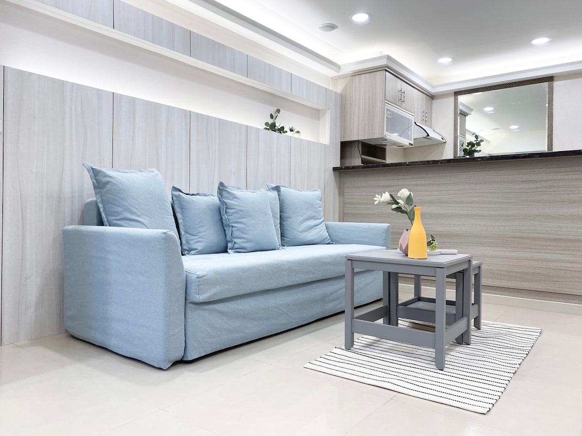 A light blue sofa is situated in a modern living area, complemented by decorative cushions. A small gray coffee table stands in front, holding a flower vase. The space features a sleek kitchen in the background with matching cabinetry.