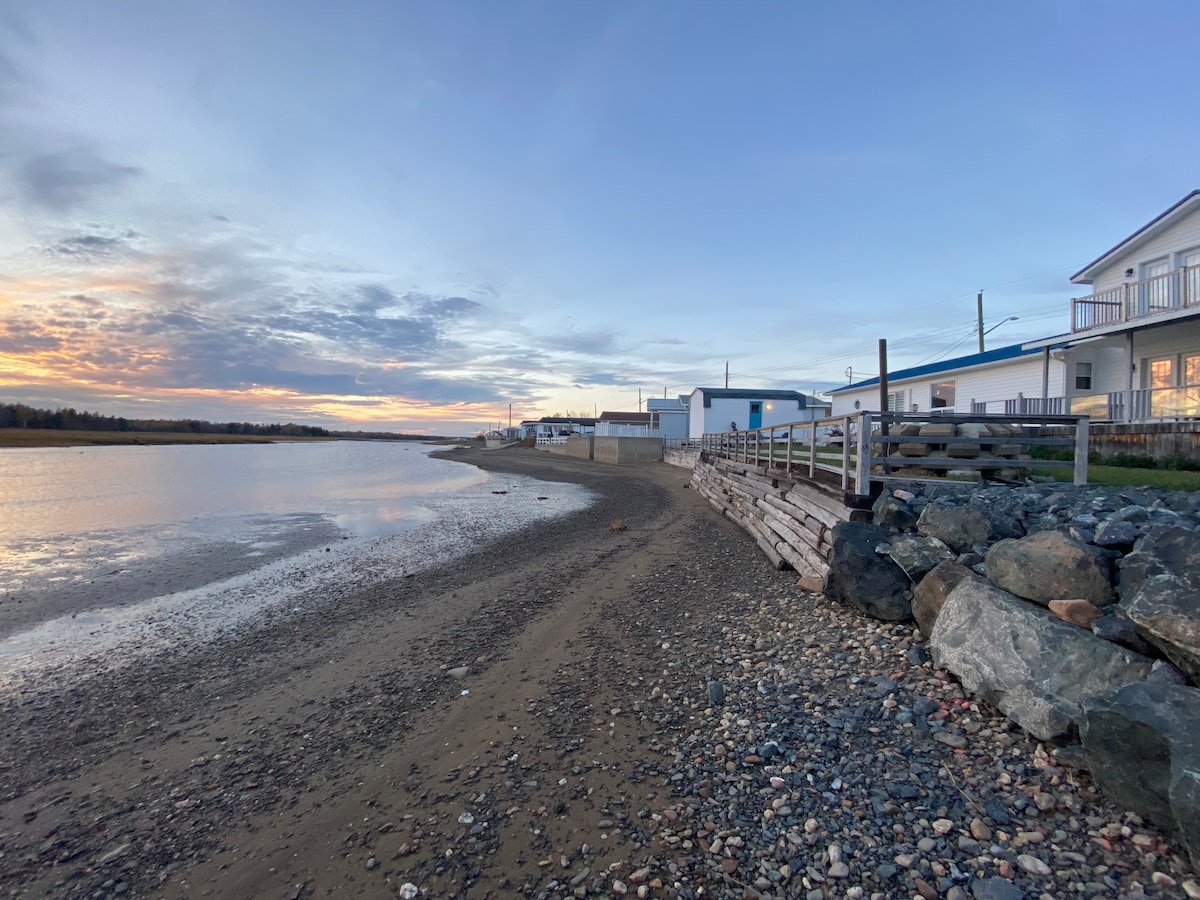 A calm shoreline is depicted with pebbles and sand, extending toward the horizon. Buildings are visible in the distance, while the water reflects the colors of the sunset. A wooden walkway runs alongside the beach, bordered by large rocks.