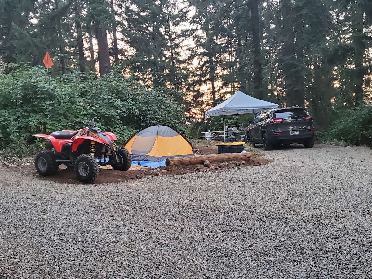 An outdoor camping site is depicted, featuring a blue and orange tent alongside a parked vehicle. An all-terrain vehicle is positioned nearby, with a canopy providing shaded space. Lush greenery surrounds the area, and gravel covers the ground.