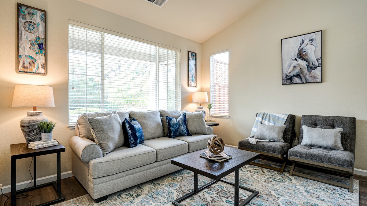 A comfortable living area features a spacious gray sofa adorned with decorative pillows. Natural light filters through large windows, illuminating the space. A coffee table sits centrally, complemented by stylish accent chairs and artwork on the walls, creating a welcoming atmosphere.