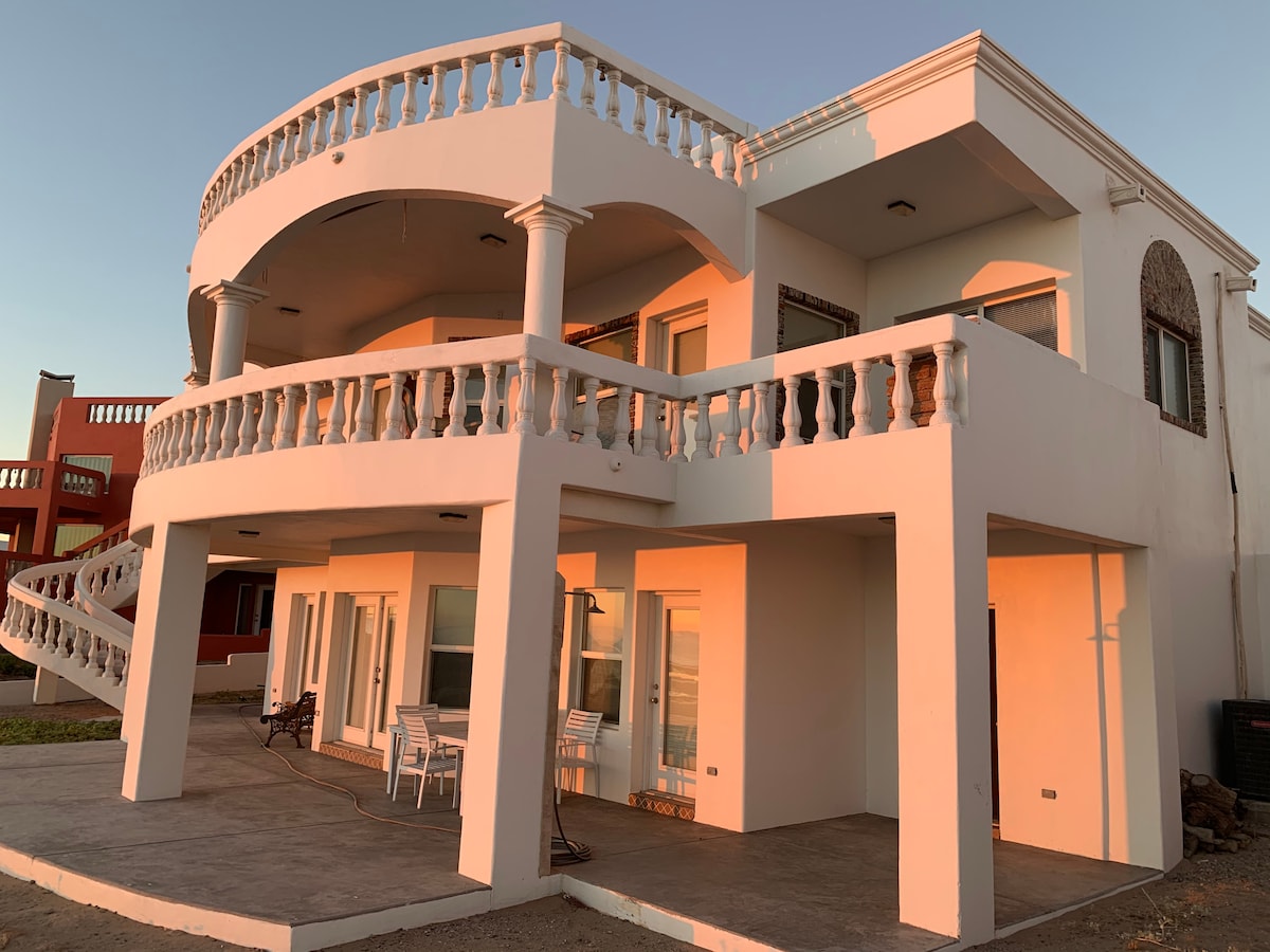 The exterior of a two-story house is featured, showcasing a spacious terrace with decorative railings. Large windows allow natural light to fill the interior spaces. A patio area is visible, complemented by outdoor furniture, creating an inviting setting for relaxation.
