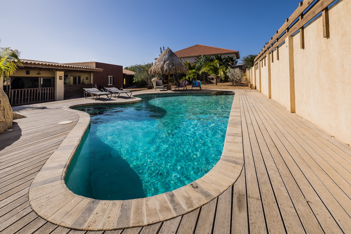 A clear blue swimming pool is outlined by wooden decking, with lounge chairs positioned nearby. The surrounding area includes a thatched roof structure, palm trees, and warm earth-toned buildings, contributing to a relaxed outdoor setting.
