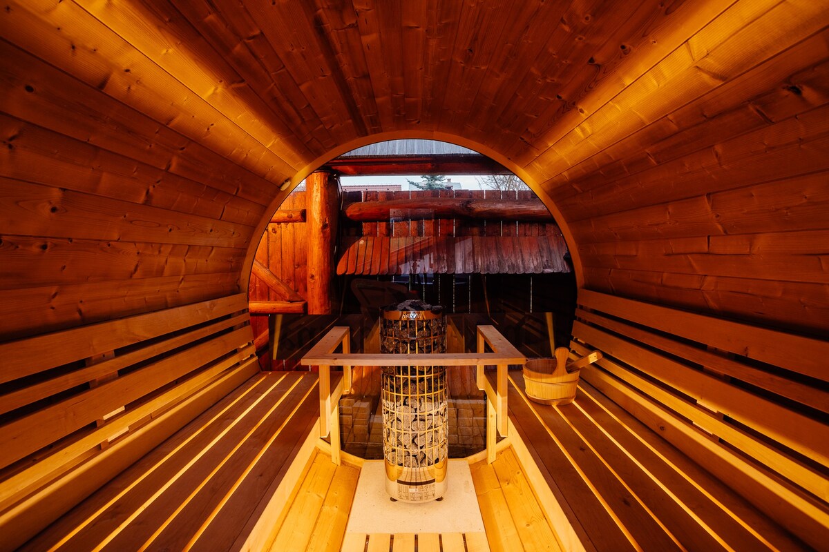 Inside the wellness area, warm wooden interiors create a calming environment. Wooden benches line the walls, forming an inviting semicircular seating arrangement. A central wooden barrel adds a rustic touch, illuminated by soft lighting that enhances the tranquil atmosphere.