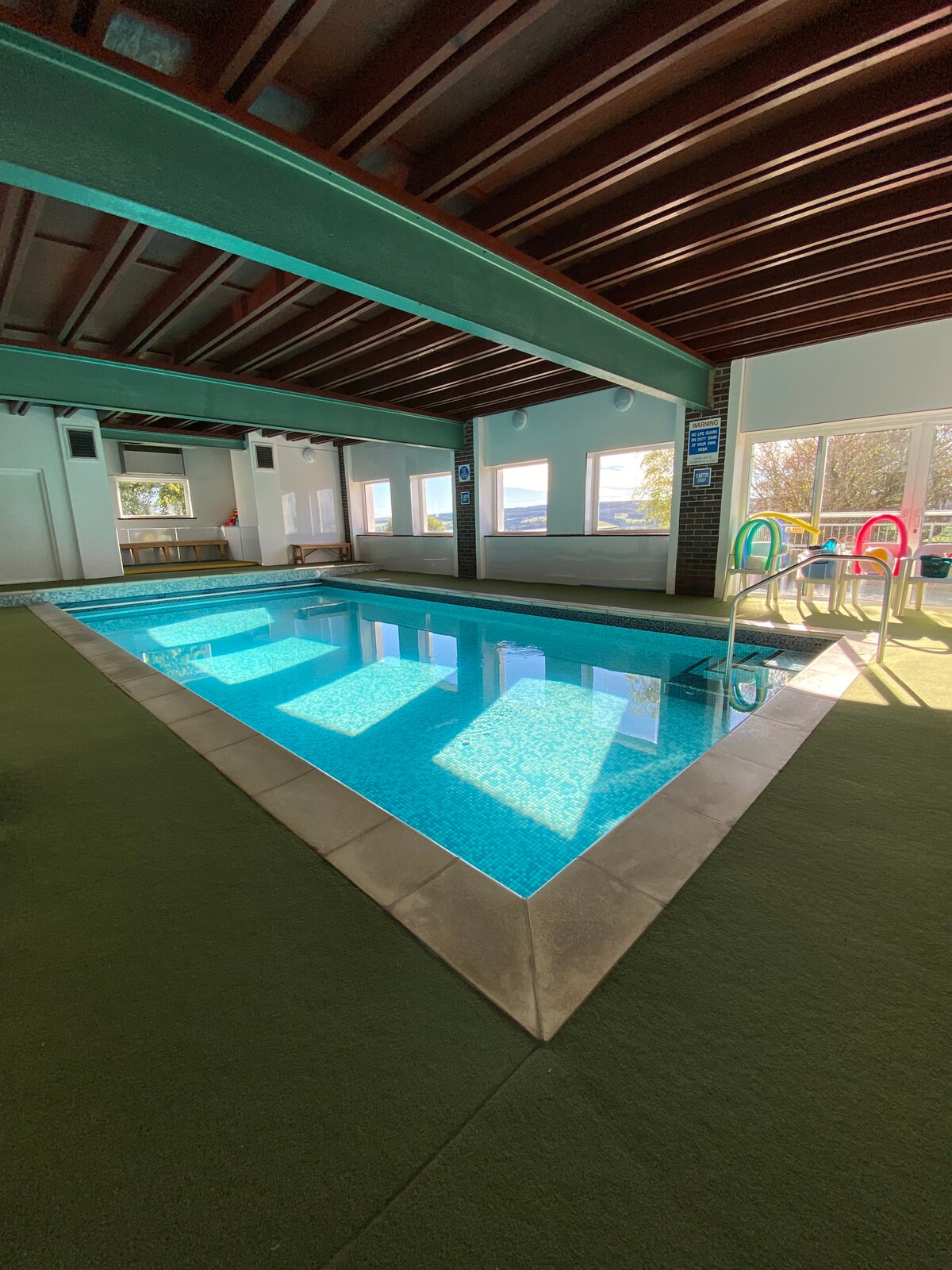 An indoor swimming pool is framed by large glass windows that allow natural light to reflect off the water’s surface. Bright green poolside chairs are positioned near the shallow end, and the surrounding area features a soft green carpet, creating a serene environment.