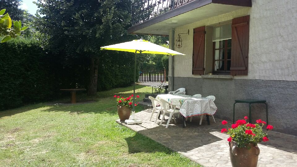 A private terrace features a table set with chairs under a bright umbrella. Flower pots with red blooms adorn the space, creating a welcoming outdoor area. Lush greenery surrounds the patio, enhancing the serene atmosphere of the garden.