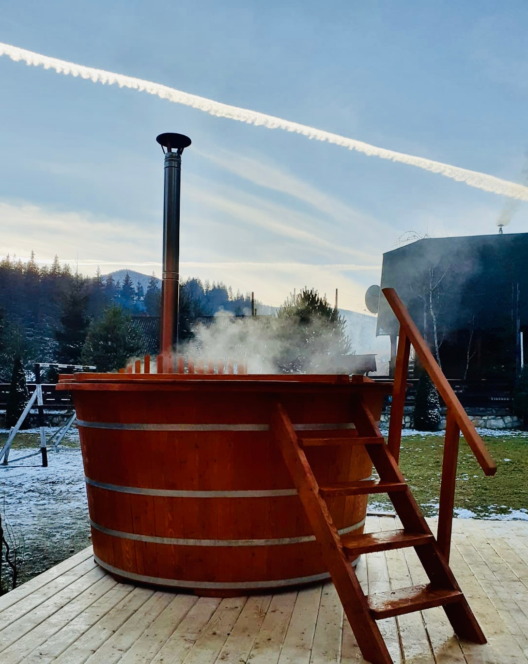 A wooden hot tub is situated on a deck, emitting steam into the chilly air. A ladder leads to the tub, which is surrounded by snow-covered greenery. In the background, rolling hills and a clear sky add to the serene setting.