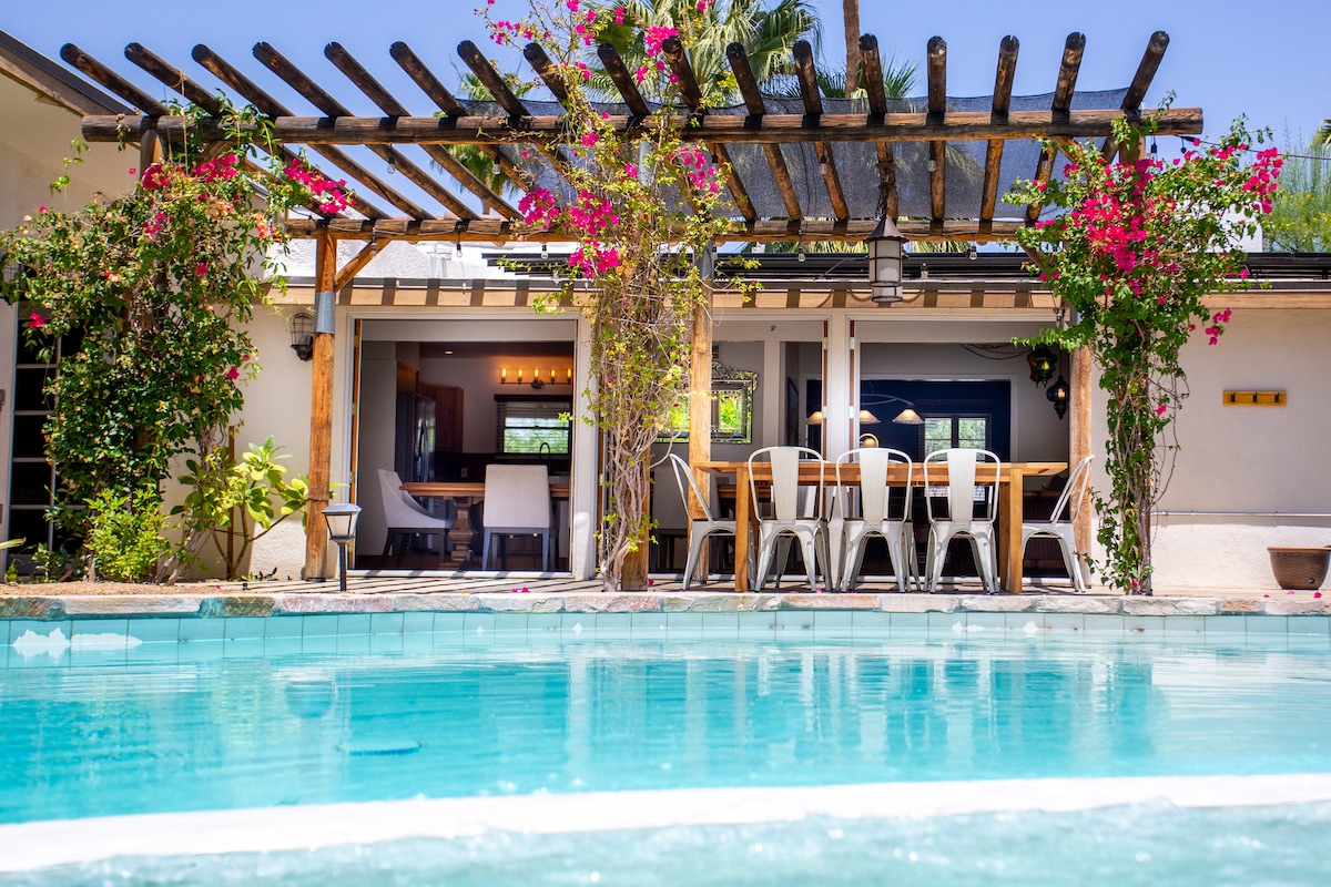 The outdoor dining area is visible under a pergola, framed by vibrant bougainvillea. A spacious table with multiple chairs awaits guests, while a sparkling saltwater pool reflects sunlight nearby. The interior of the home is glimpsed through the open doors, showcasing a cozy and inviting ambiance.
