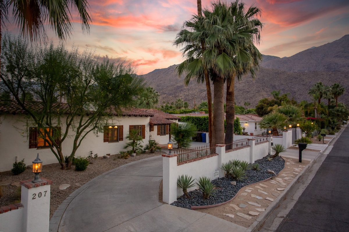 The exterior of the classic Hacienda-style home features a large driveway bordered by desert landscaping, including palm trees and low shrubs. The warm glow of outdoor lights illuminates the entrance, while the backdrop of majestic mountains and a colorful sunset sky completes the serene setting.