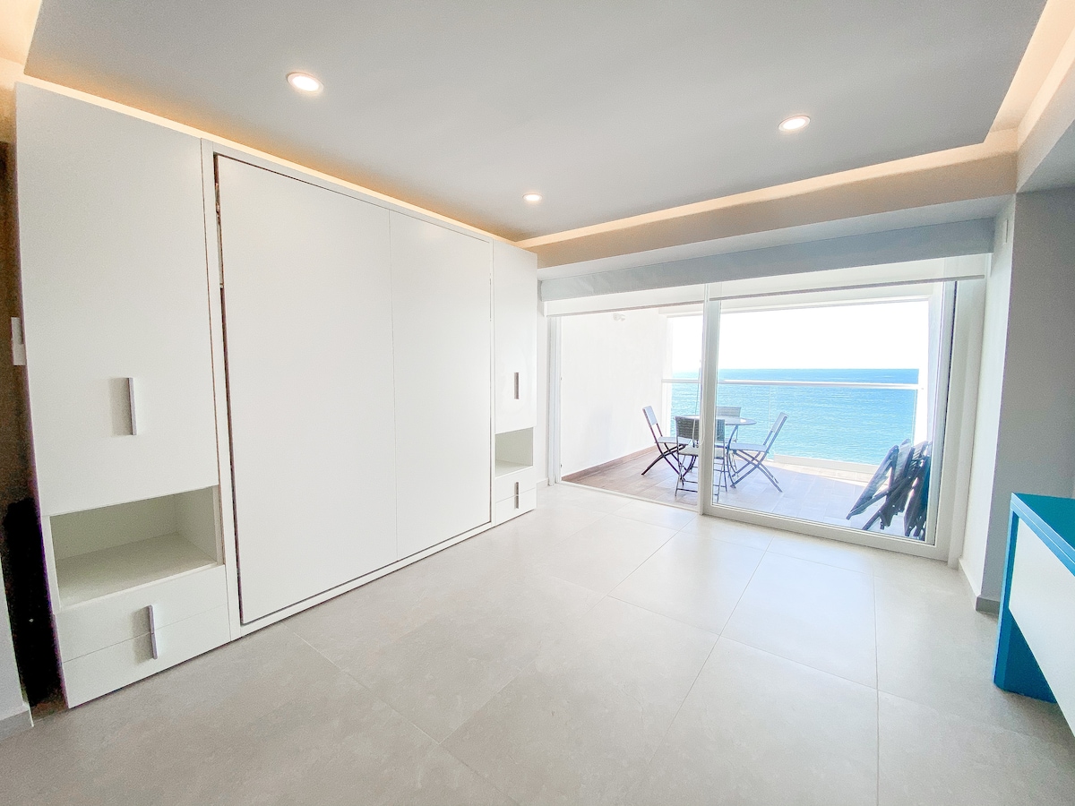 A bright living space features a sleek white built-in cabinet along one wall, with a view of the ocean visible through expansive sliding glass doors. A small outdoor dining area is set on a terrace, complete with chairs and a table.