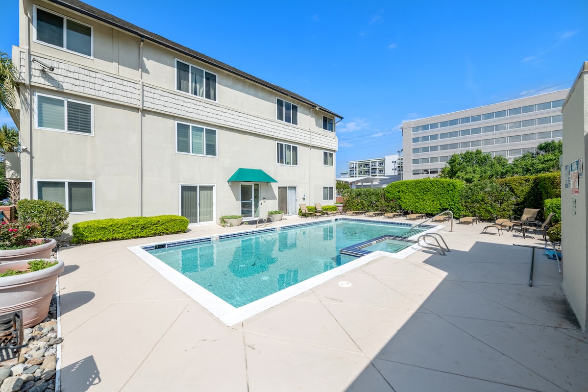 A well-maintained outdoor swimming pool is surrounded by a concrete patio, featuring lounge chairs and vibrant greenery. The three-story building has several windows and balconies, offering a bright and open environment under a clear blue sky.