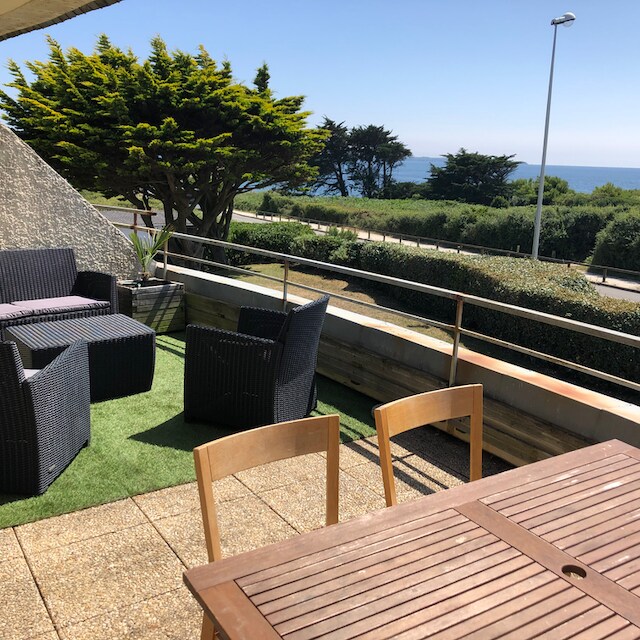 An outdoor terrace offers a view of the sea and lush greenery. Comfortable seating arrangement with dark wicker chairs surrounds a wooden table. The flooring features a textured surface, and the space is enveloped by low-maintenance plants, creating a relaxed environment.