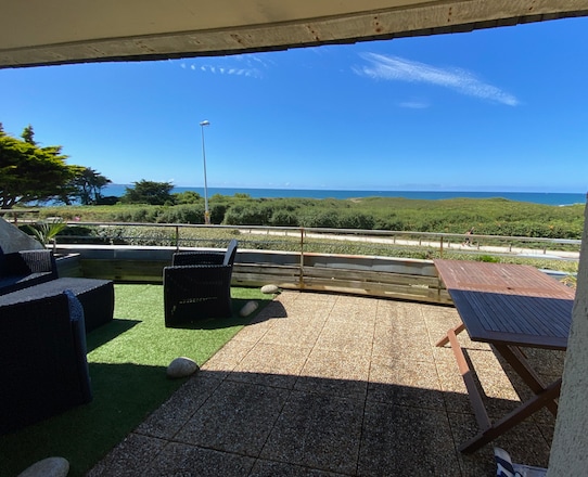 An outdoor terrace is visible, featuring a dining table and lounge seating. Green grass and decorative stones embellish the space. The sea and coastal landscape can be seen in the distance under a clear blue sky.