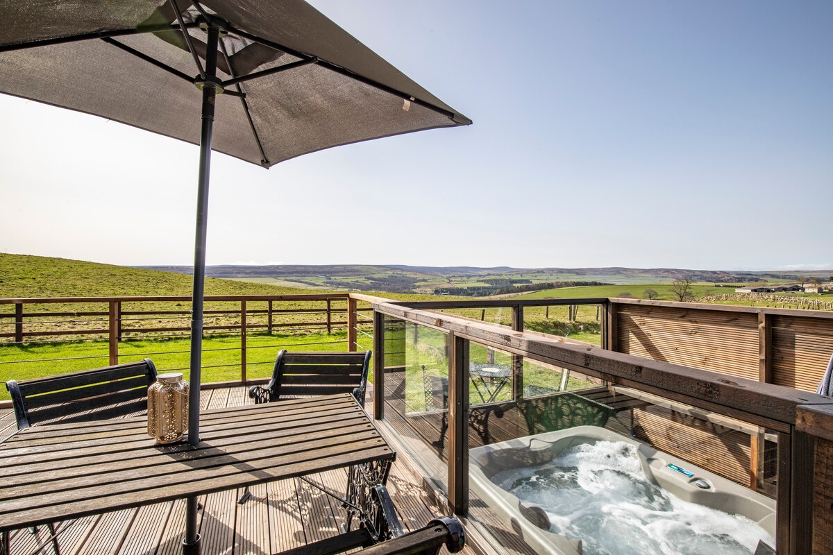An outdoor deck area features a hot tub alongside a wooden dining table and chairs, shaded by a large umbrella. The scenic view of rolling hills and distant moors is visible in the background, creating a serene environment for relaxation.