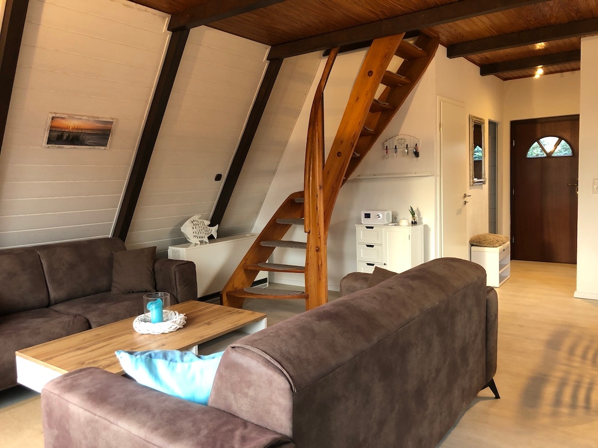 A cozy living area features two spacious sofas positioned around a light wood coffee table. A staircase leads to the upper level, while a sideboard and a window provide additional elements to the space. Soft lighting enhances the welcoming ambiance.