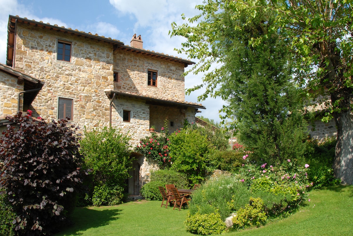 The exterior of the rustic stone building is surrounded by lush greenery and blooming flowers, creating a serene outdoor space. A seating area with wooden chairs and a small table is positioned on the lawn, inviting relaxation in the garden.