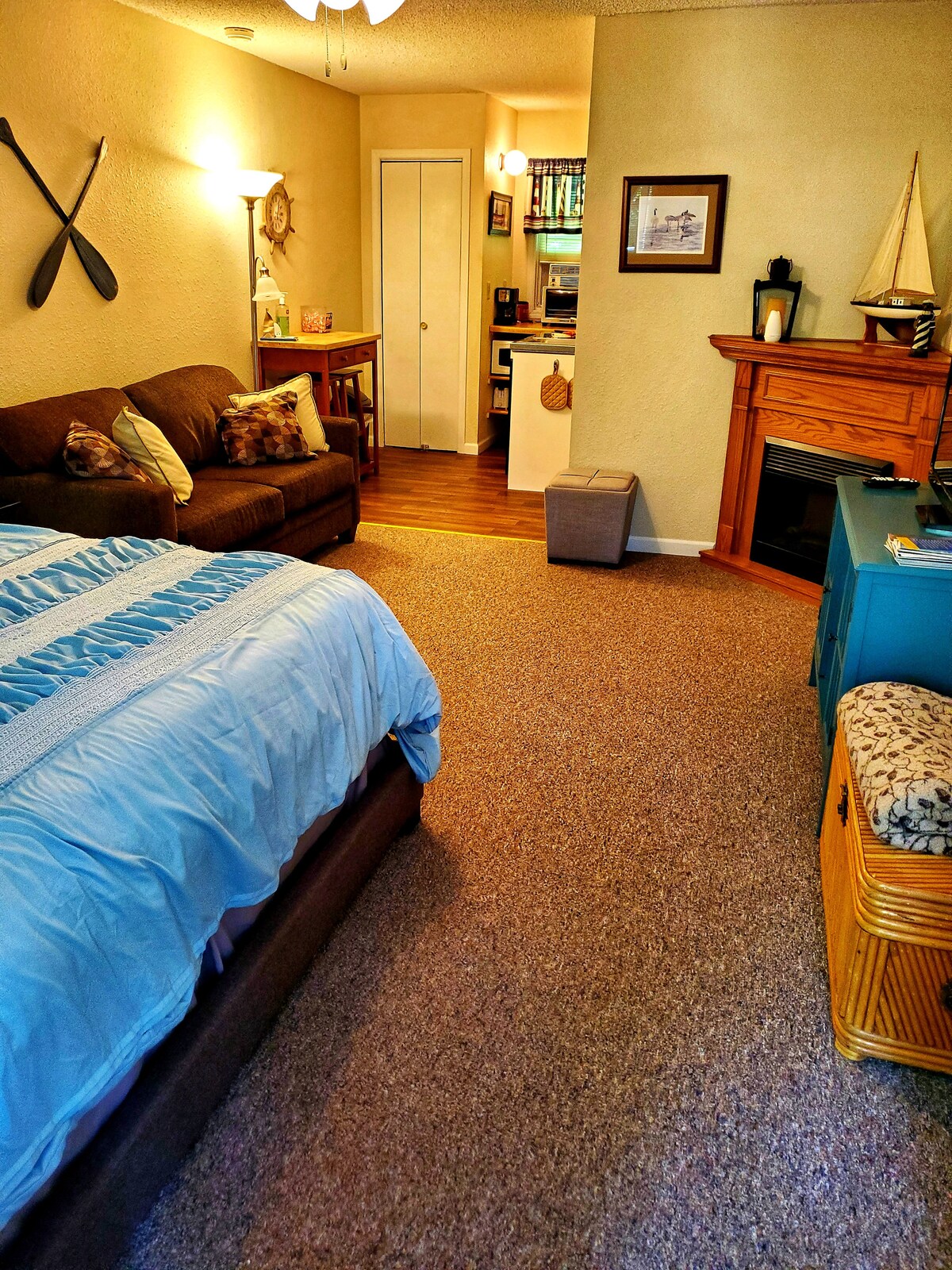 A spacious studio layout features a cozy bed dressed in light blue linens, accompanied by a soft couch and small dining area. Nearby, the kitchen is visible with wooden accents. A decorative fireplace adds a subtle charm, and soft lighting enhances the inviting environment.