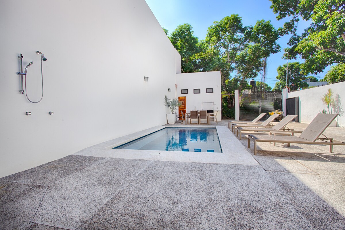 A swimming pool is framed by light-colored lounge chairs, positioned on the textured patio. A shower is mounted on the wall beside the pool. Lush greenery and trees provide shade in the background, contributing to the outdoor space's inviting atmosphere.