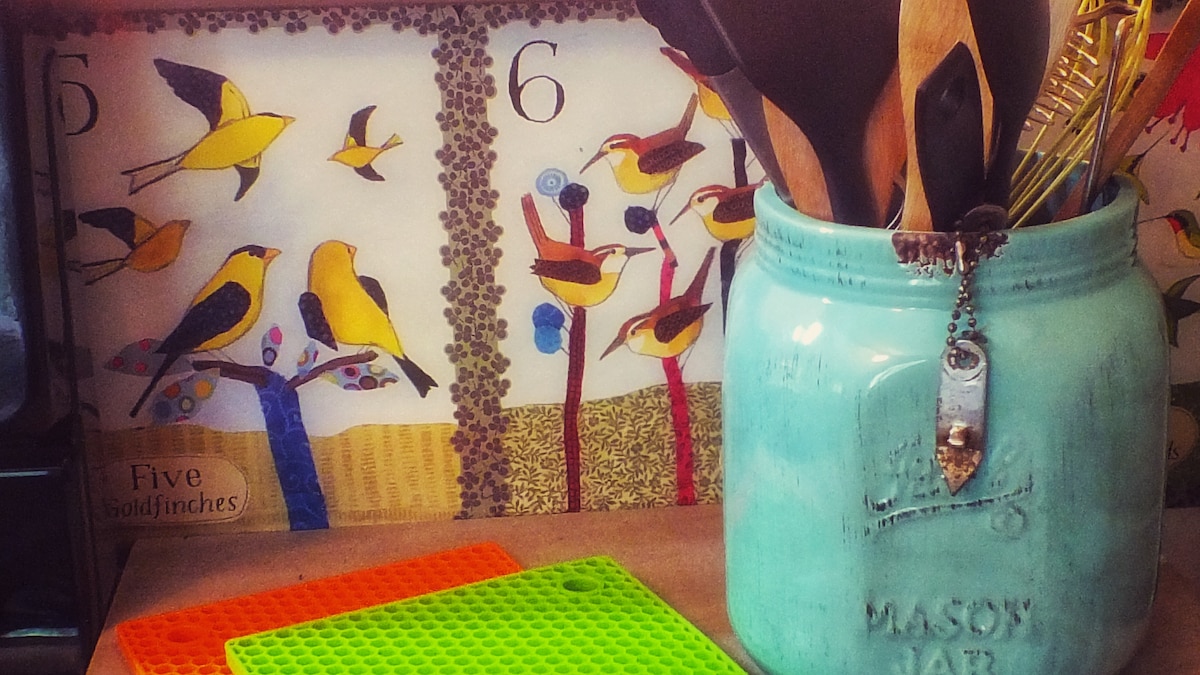 A vibrant kitchen area displays a colorful bird-themed poster in the background. A mason jar in a turquoise hue holds various cooking utensils. Two silicone mats in orange and green are placed on the countertop, adding a playful touch to the space.