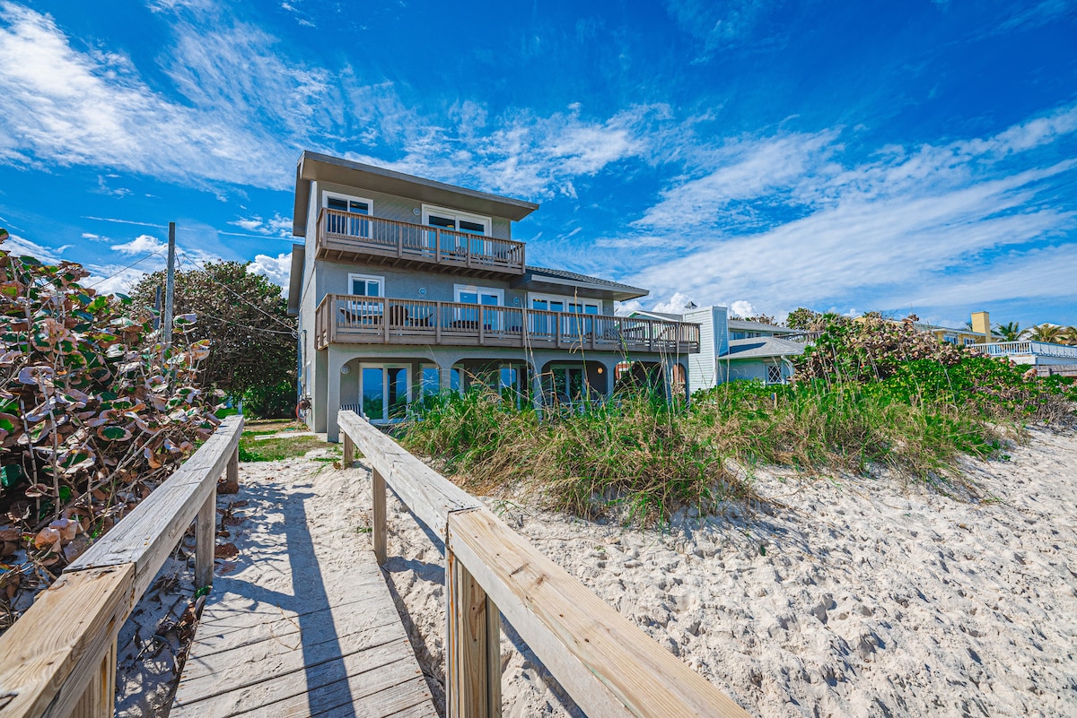 A three-story coastal home is shown, surrounded by lush greenery and sandy pathways leading to the beach. Balconies extend from the second and third floors, offering views of the ocean. Blue skies with scattered clouds frame the structure, creating a serene beachside atmosphere.