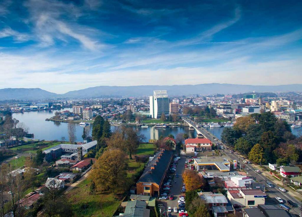 The aerial view captures the vibrant city of Valdivia, showcasing a mix of residential buildings and natural landscapes. The calm river winds through the scenery, with the town's modern architecture in the background and distant mountains under a clear sky.