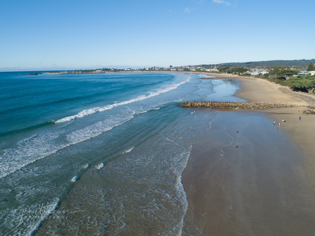 A panoramic view of a sandy beach meeting the turquoise ocean, capturing gentle waves lapping at the shore. Rocky formations are visible in the distance, with people walking along the water's edge. A clear blue sky provides a bright backdrop to the serene coastal scene.