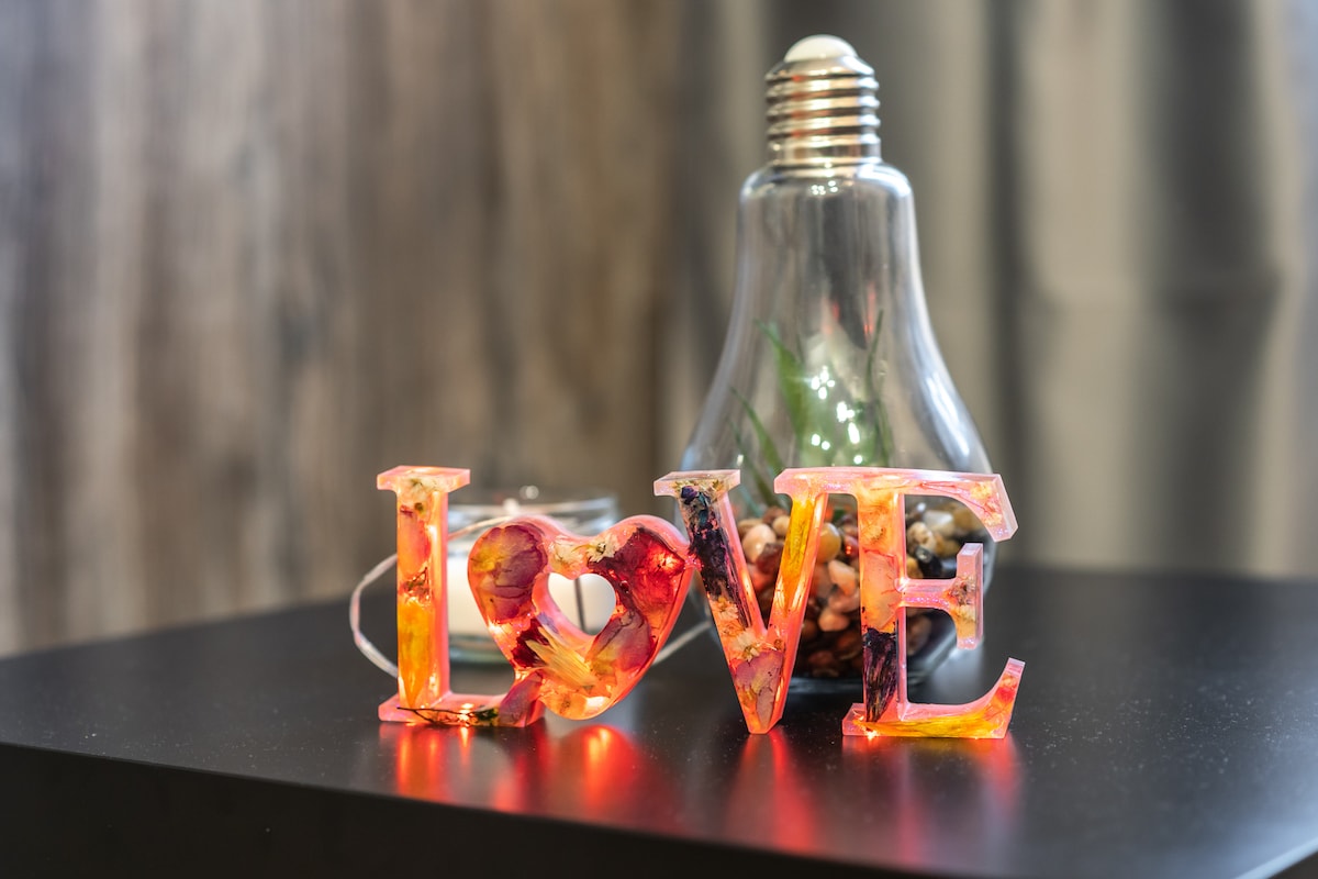 A decorative arrangement features a colorful 'LOVE' sculpture in front of a glass lamp. The lamp showcases decorative stones inside, contributing to a warm ambiance. The focus is on the vibrant hues of the sculpture, accentuated by ambient lighting.