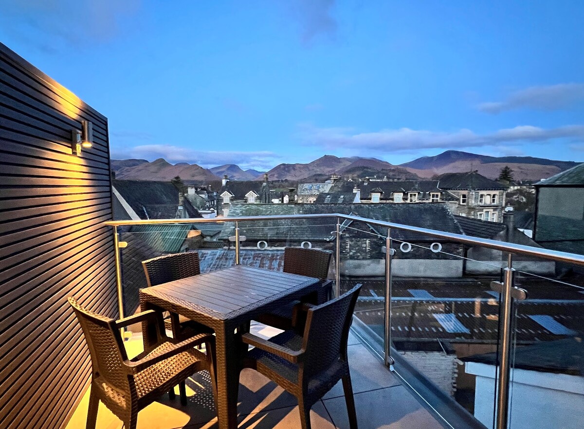 A roof terrace features a wooden table surrounded by four sleek wicker chairs, offering panoramic views of the surrounding fells and town below. Soft lighting enhances the evening ambiance, with a clear sky and distant mountains visible in the background.