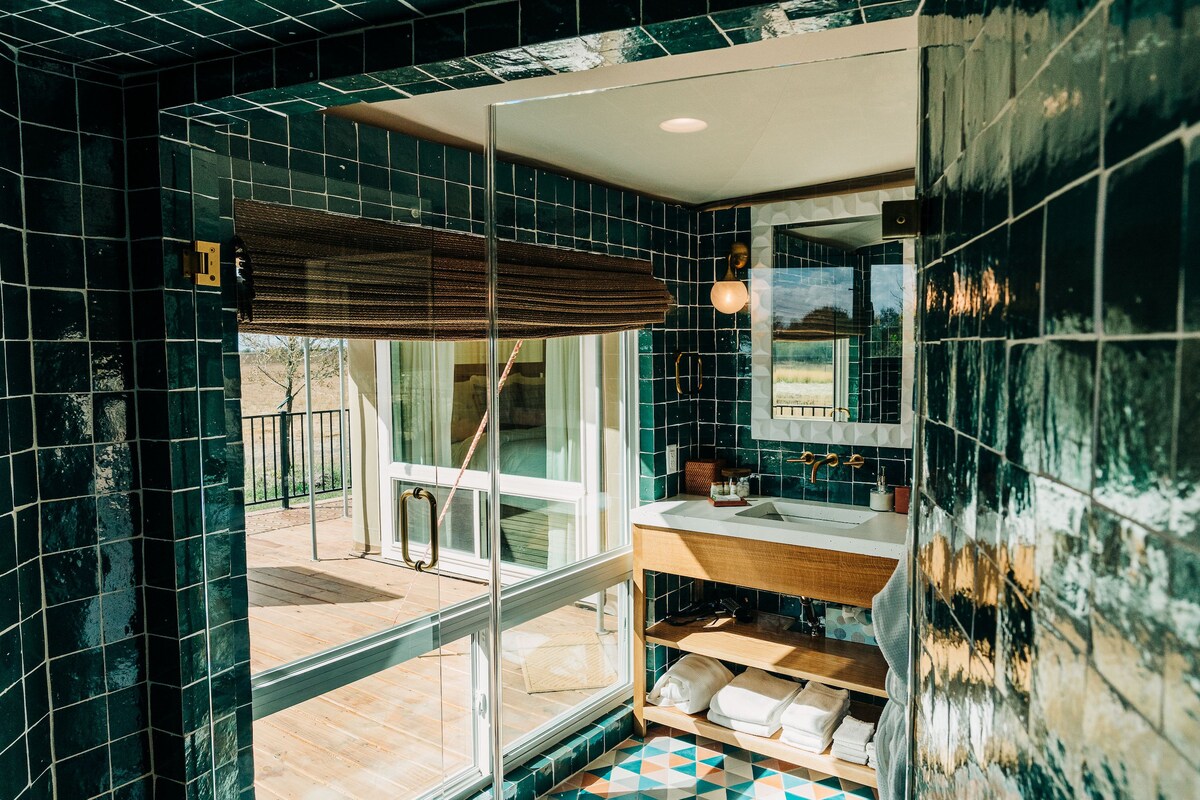 A modern bathroom features vibrant green tiles covering the walls. A large shower is visible, alongside a spacious vanity with a sink and wooden accents. Soft white towels are neatly arranged nearby. Natural light enters through a glass door leading to the private deck.