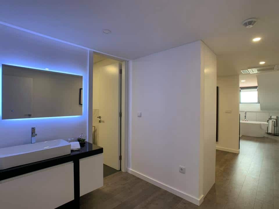 A modern bathroom is presented, featuring a sleek vanity with a large mirror illuminated by soft blue LED lighting. The space is designed with minimalist decor, highlighted by light-colored walls and a wooden floor, leading to a separate area visible in the background.