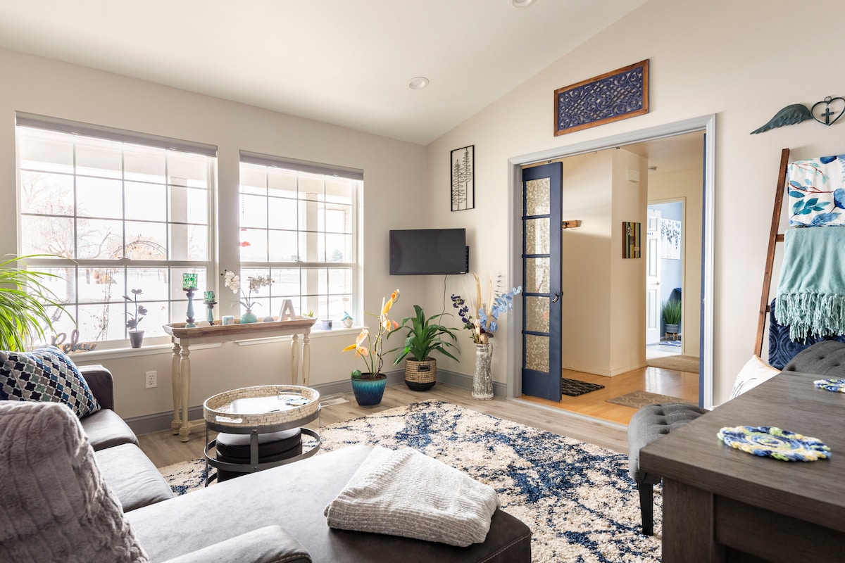 A bright living area is showcased with large windows allowing natural light to fill the space. A cozy seating arrangement is complemented by a decorative rug. An entry door with glass panels enhances the openness, while plants and decorative elements add to the inviting ambiance.
