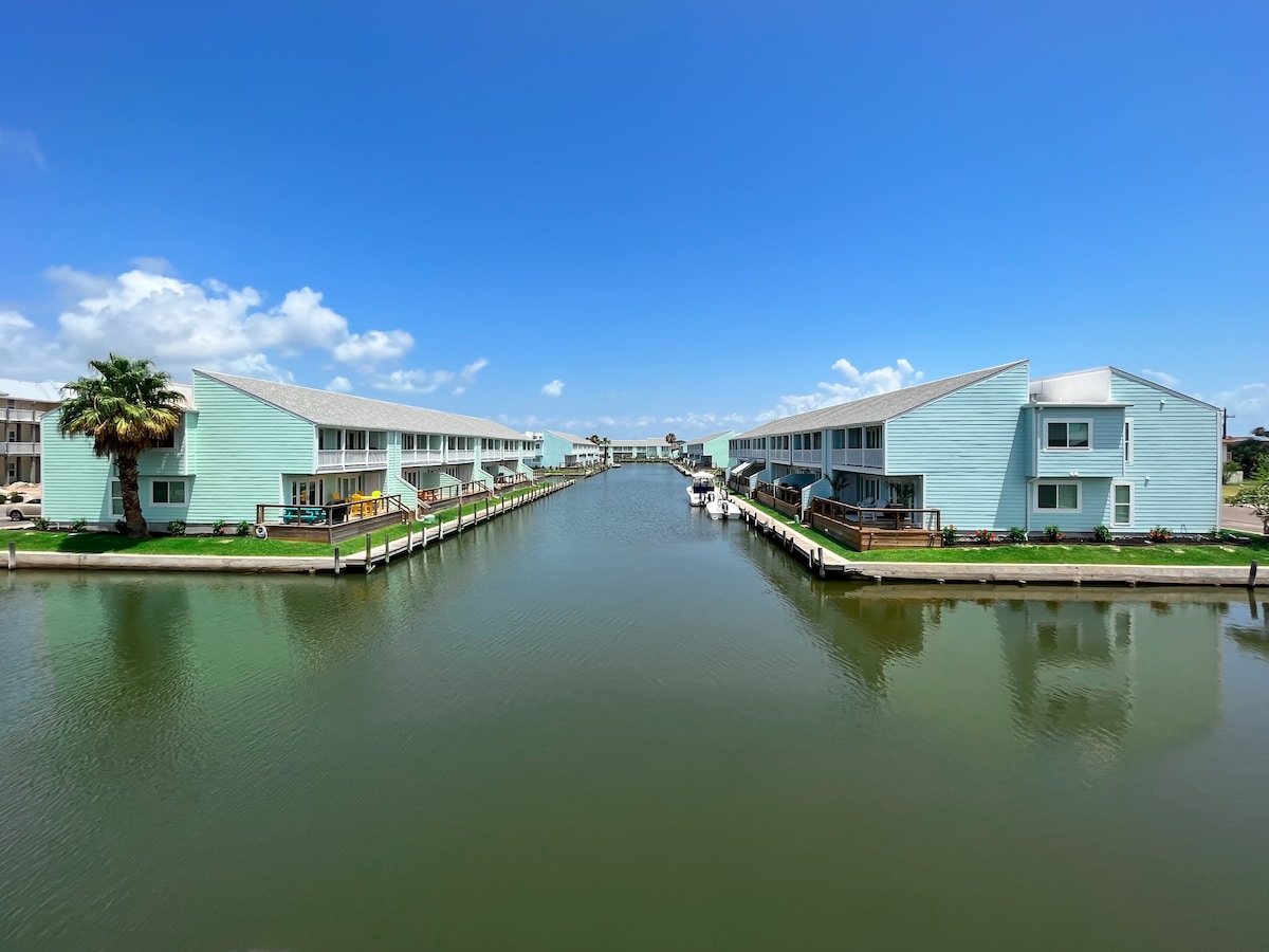 A serene waterfront view showcases two adjacent buildings with light blue exteriors, reflecting the sun against a clear blue sky. A calm canal runs between the buildings, with docks lining either side, creating a welcoming environment for boat access.